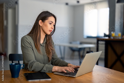 Serious dedicated businesswoman using laptop sitting at the table in a home office  looking at device screen  communicating online  writing emails  distantly working or studying on computer at home.