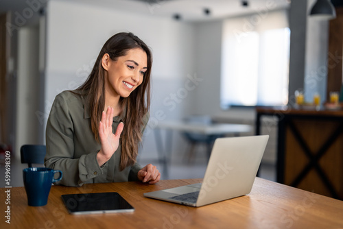 Kind smiling businesswoman, satisfied female entrepreneur waving hand looking at laptop during virtual video conference call in home office. © Dorde