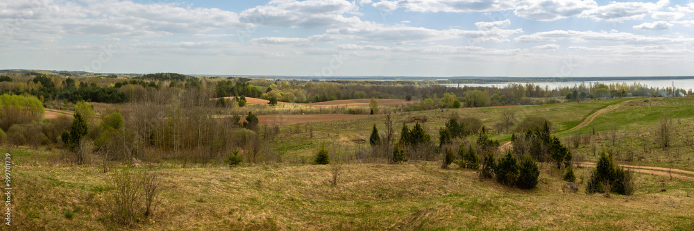 wide panoramic view of a hilly field with wild grass and trees, farmland, forest belts and a lake on a spring day under a blue cloudy sky