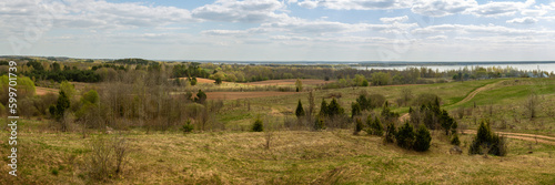 wide panoramic view of a hilly field with wild grass and trees, farmland, forest belts and a lake on a spring day under a blue cloudy sky photo