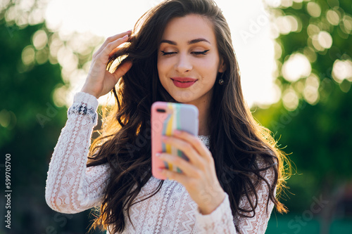 A happy young woman is standing outdoors on a sunny summer day, touching her hair and sending flirty messages.