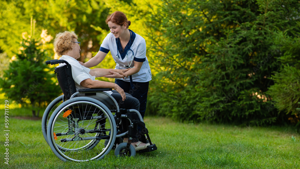 A nurse holds an elderly caucasian woman in a wheelchair by the hand as support. Nurse walks with a patient in the park. A nurse holds an elderly caucasian woman in a wheelchair by the hand as support. Nurse walks with a patient in the park.