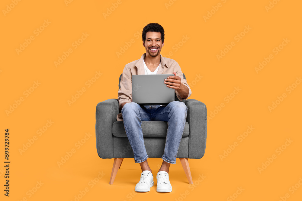 Happy man with laptop sitting in armchair on orange background