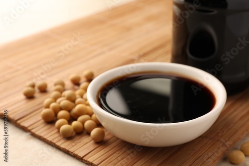 Soy sauce in bowl and soybeans on bamboo mat, closeup photo