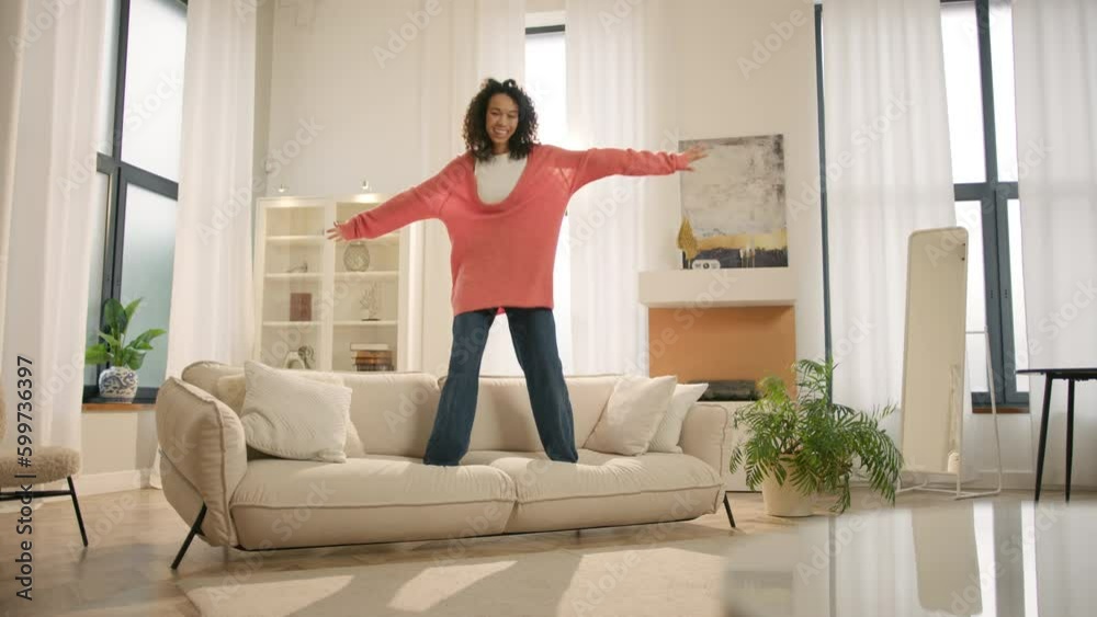 Excited happy young woman of color jumping happily on modern sofa. Afro girl homeowner having fun on move in day, celebrating mortgage approval, relocation to upgraded apartment. Bank mortgage concept Excited happy young woman of color jumping happily on modern sofa. Afro girl homeowner having fun on move in day, celebrating mortgage approval, relocation to upgraded apartment. Bank mortgage concept
