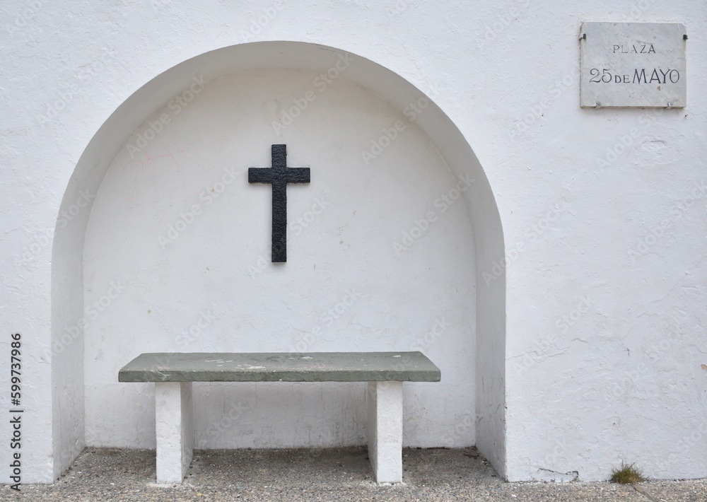 Black carved cross on a white wall with a bench in a small town square