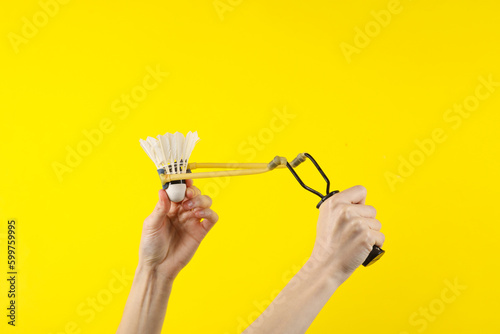Hands holding slingshot with shuttlecock on a yellow background.