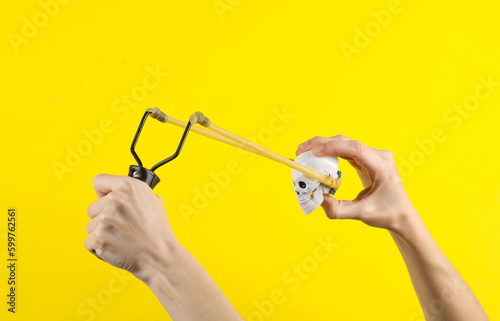 Hands holding slingshot with toy skull on a yellow background. Halloween theme, trick or treat