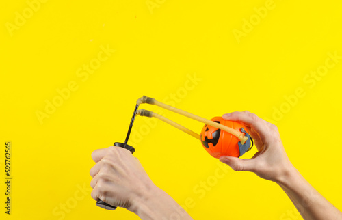 Hands holding slingshot with toy Jack pumpkin bucket on a yellow background. Halloween theme, trick or treat