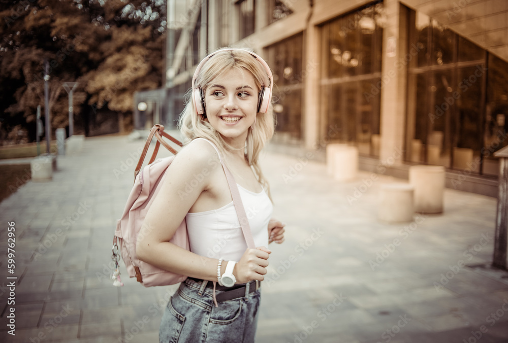 Charming cute young girl listening to music on headphones with a backpack behind her shoulders in the city