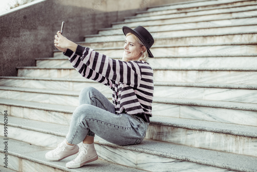 Young charming hipster girl in a hat makes a selfie on the phone while sitting on the stairs outdoors