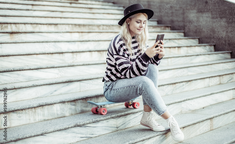Young charming hipster girl in hat using phone while sitting on the stairs with skateboard outdoors