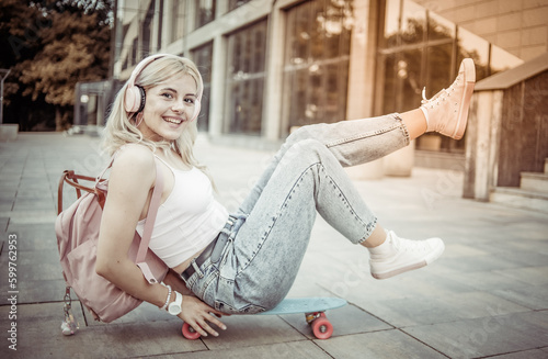 Young smiling cool girl sitting on skateboard and listen music in headphones outdoors