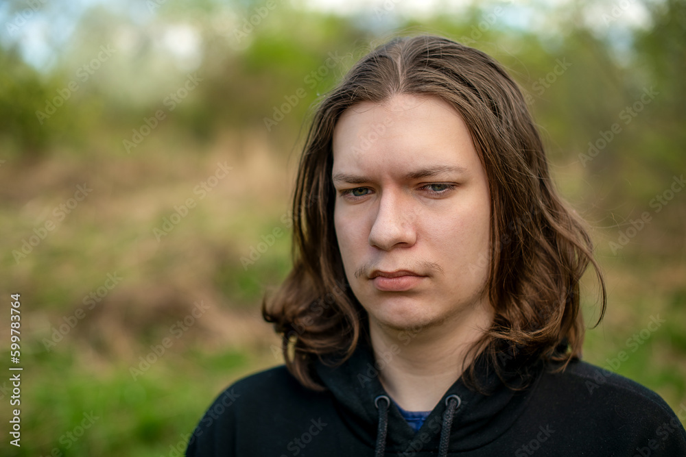 An attractive young man with long blond hair and wearing a black hoodie poses in front of the camera in nature on a clear summer day. An attractive young man with long blond hair and wearing a black hoodie poses in front of the camera in nature on a clear summer day.