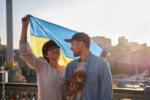 Young smiling couple with dog holding a yellow and blue flag of Ukraine on a background of Kyiv on the sunset. Glory to Ukraine High quality photo