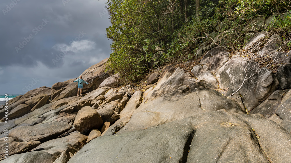 A man stands on the slope of a coastal cliff, his hands spread apart. Green plants on the rocks. Clouds in the blue sky. The ocean is visible in the distance. Seychelles. Moyenne Island. 