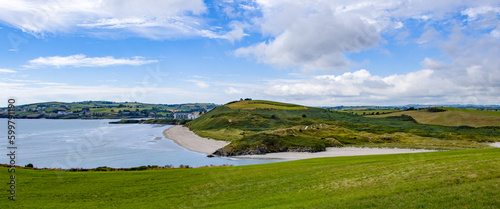 The green hills of Inchydoney Island and the famous Irish beach on a sunny summer day. Clear sky with white clouds over the Irish coast. Irish summer landscape. Green grass field near water