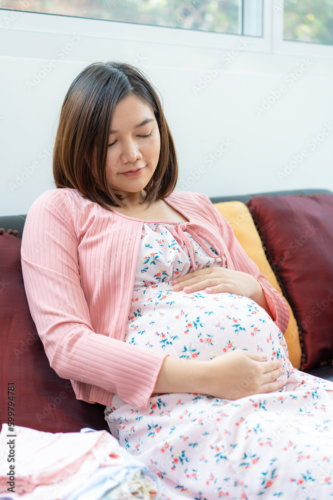 Happy young pregnant Asian woman in maternity clothes sitting on a sofa in her living room holding her belly Happy young pregnant Asian woman in maternity clothes sitting on a sofa in her living room holding her belly