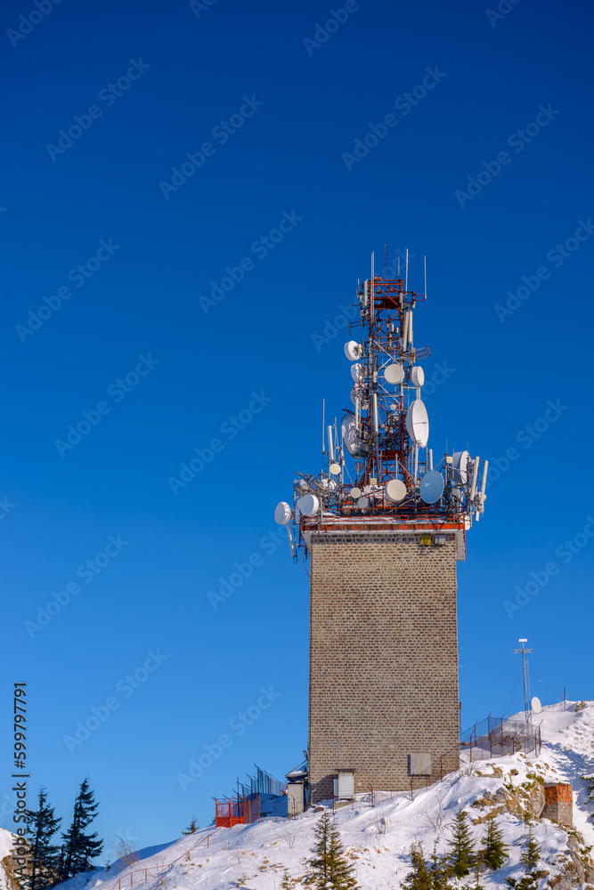 Naklejka premium Tower of telecommunication with many antennas, in a beautiful winter mountains landscape, with trees covered in snow and copy space.