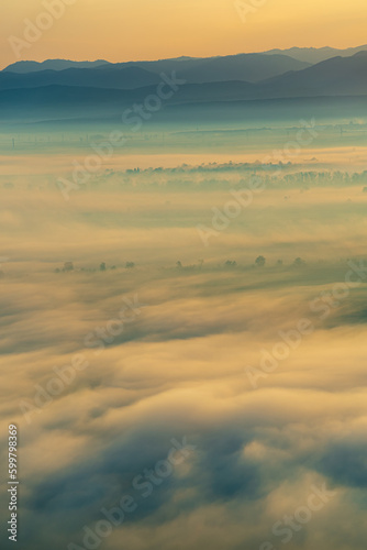 Aerial view of the valley in early morning mist, beautiful in the highlands. Low clouds and fog cover the sleeping meadow. Alpine mountain valley mists landscape at dawn. Serene moment in rural area