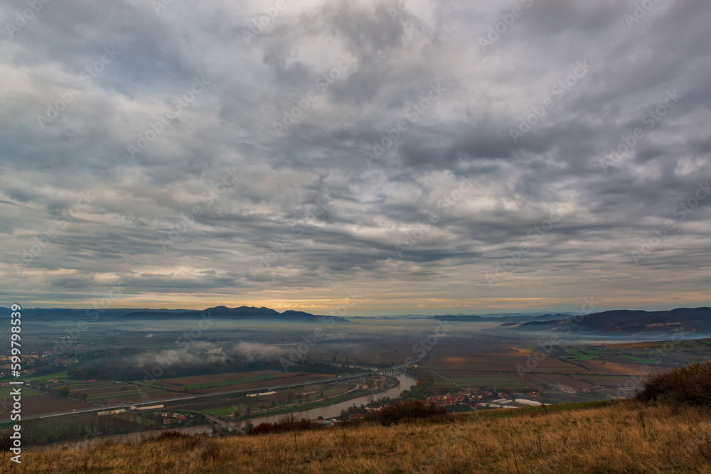 Serene moment in rural area in beautiful early morning mist. Panoramic view of a sleeping meadow valley covered with low clouds and fog, a highway crossing a river, rolling hills and villages