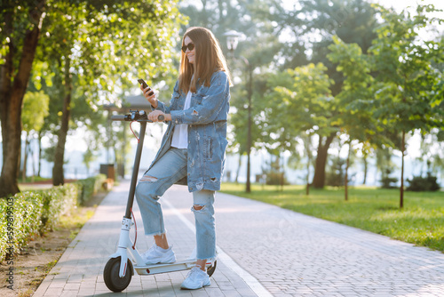 Young female legs on an electric scooter. Beautiful woman rides an electric scooter in the park on a sunny day. Ecological transport. Active lifestyle.