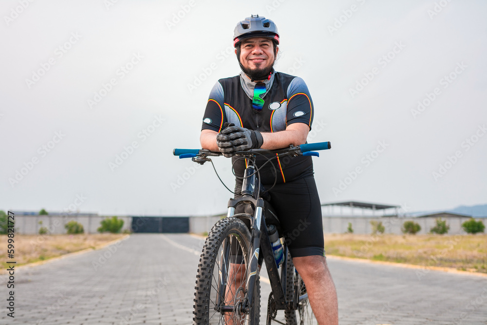 Fototapeta premium Smiling cyclist on his bike in sports outfit. Chubby male cyclist in sportswear riding a bicycle outdoors