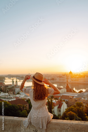 Beautiful woman in hat enjoys stunning view of the city at dawn. Back view. Tourist is looking at landscape of city from height. Travel in Europe.