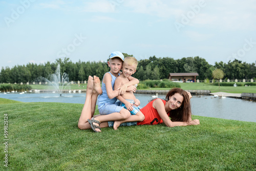 Happy mom and sons. Mom and children. Boys brothers sit on the back of their mother, who lies on the green grass against the backdrop of a beautiful summer landscape in the park in summer.