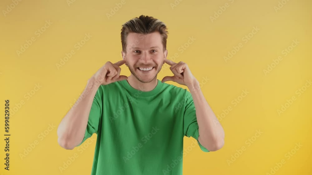 Smiling man posing for the camera with his fingers plugging his ears. A guy in a green T-shirt stands against a yellow wall and pretends not to listen. Smiling man posing for the camera with his fingers plugging his ears. A guy in a green T-shirt stands against a yellow wall and pretends not to listen.