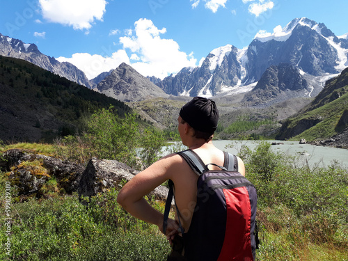 A Caucasian man without a T-shirt with a backpack stands back and looks at high snowy mountains and a mountain lake. The concept of tourism and travel.