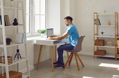 Serious focused businessman or freelancer working with laptop at home office. Young man in casual clothes sitting at desk working on project, researching, making notes on paper