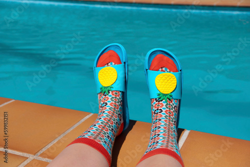 Feet with colorful socks and blue pool sandals at the edge of a swimming pool. Summer concept. Aesthetic summer