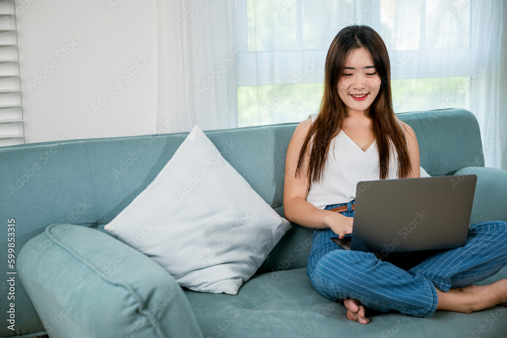 Happy woman typing email on notebook computer, Asian young female smiling sitting relaxing on sofa using laptop in living room at home, freelance browsing through the internet during free time Happy woman typing email on notebook computer, Asian young female smiling sitting relaxing on sofa using laptop in living room at home, freelance browsing through the internet during free time