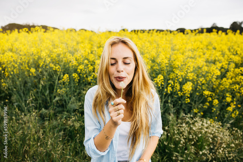 Beautiful young blonde cheerful woman, blowing dandelion seeds amidst a field of blooming yellow rapeseed flowers