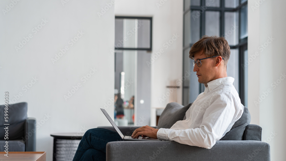 Businessman side view typing in laptop sitting on sofa in modern apartment Businessman side view typing in laptop sitting on sofa in modern apartment