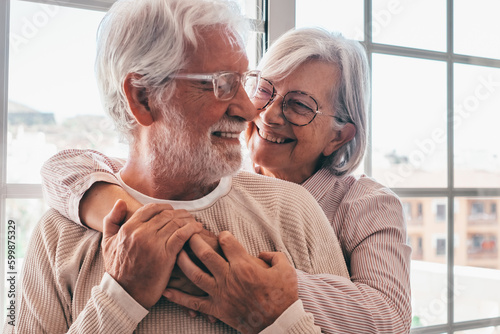 Smiling senior family couple of Caucasian people hugging in front the window, elderly man and woman with glasses embracing each other in a moment of tendress
