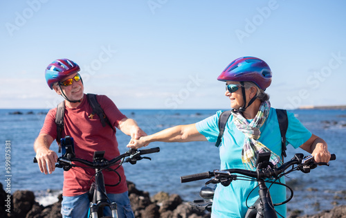Cheerful senior couple enjoying riding bikes together at sea to be fit and healthy. Active seniors wearing helmets having fun training in outdoors. Authentic elderly retired life concept