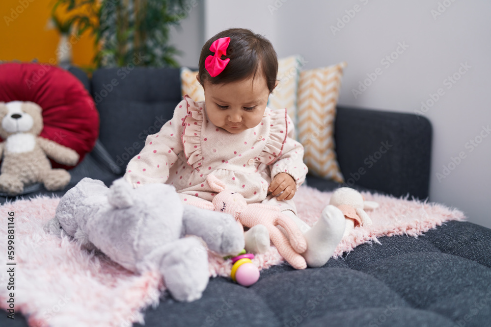Fototapeta premium Adorable hispanic baby playing with doll sitting on sofa at home
