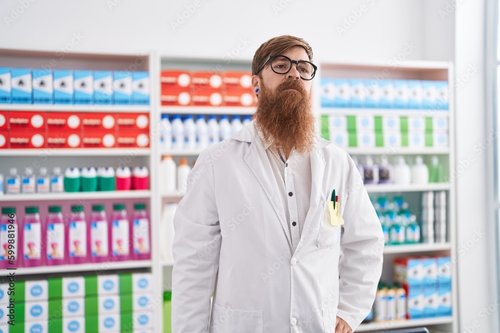 Young redhead man pharmacist standing with serious expression at pharmacy