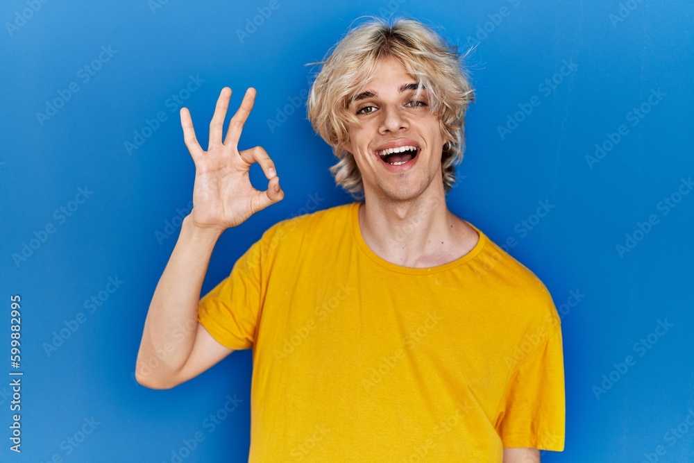 Young modern man standing over blue background smiling positive doing ok sign with hand and fingers. successful expression. Young modern man standing over blue background smiling positive doing ok sign with hand and fingers. successful expression.