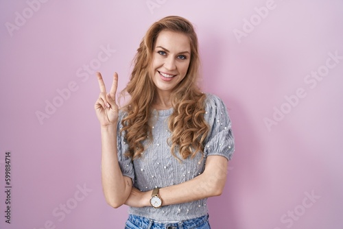 Beautiful blonde woman standing over pink background smiling with happy face winking at the camera doing victory sign with fingers. number two. © Krakenimages.com