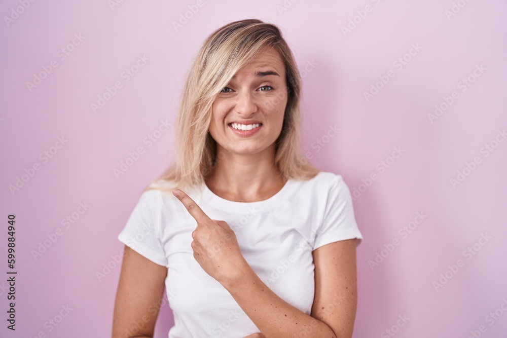 Young blonde woman standing over pink background pointing aside worried and nervous with forefinger, concerned and surprised expression Young blonde woman standing over pink background pointing aside worried and nervous with forefinger, concerned and surprised expression