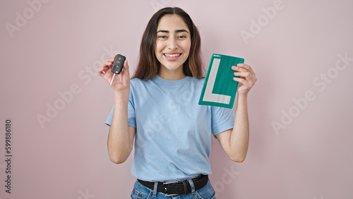 Young beautiful hispanic woman smiling confident holding key of new car and amateur driver license over isolated pink background