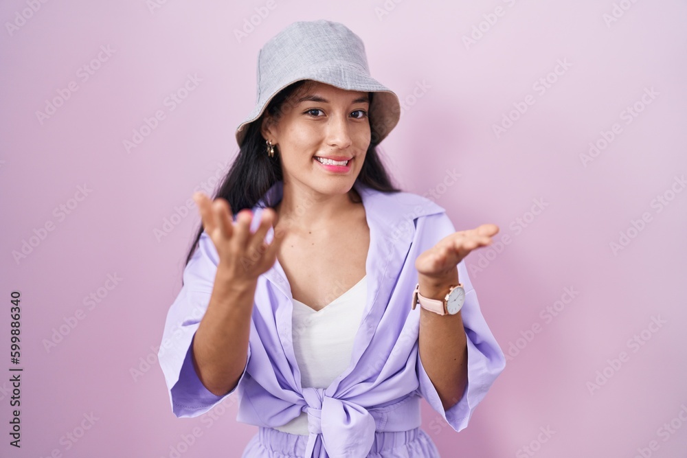 Young hispanic woman standing over pink background wearing hat smiling cheerful offering hands giving assistance and acceptance. Young hispanic woman standing over pink background wearing hat smiling cheerful offering hands giving assistance and acceptance.