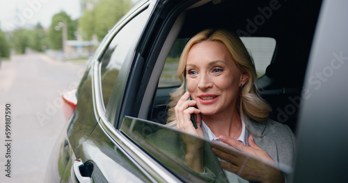 Beautiful adult caucasian woman rides on rear passenger seat of car, looking wonder out of the window during important phone call