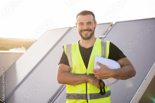 A specialized technician with protective clothing is posing smiling while looking at the camera in front of some solar panels to heat water from some houses.Concept of solar panels, renewable energy photo