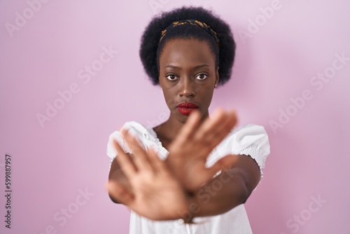 African woman with curly hair standing over pink background rejection expression crossing arms and palms doing negative sign, angry face