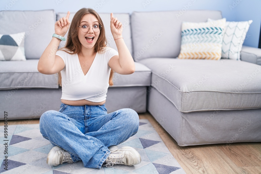Young caucasian woman sitting on the floor at the living room smiling amazed and surprised and pointing up with fingers and raised arms. Young caucasian woman sitting on the floor at the living room smiling amazed and surprised and pointing up with fingers and raised arms.