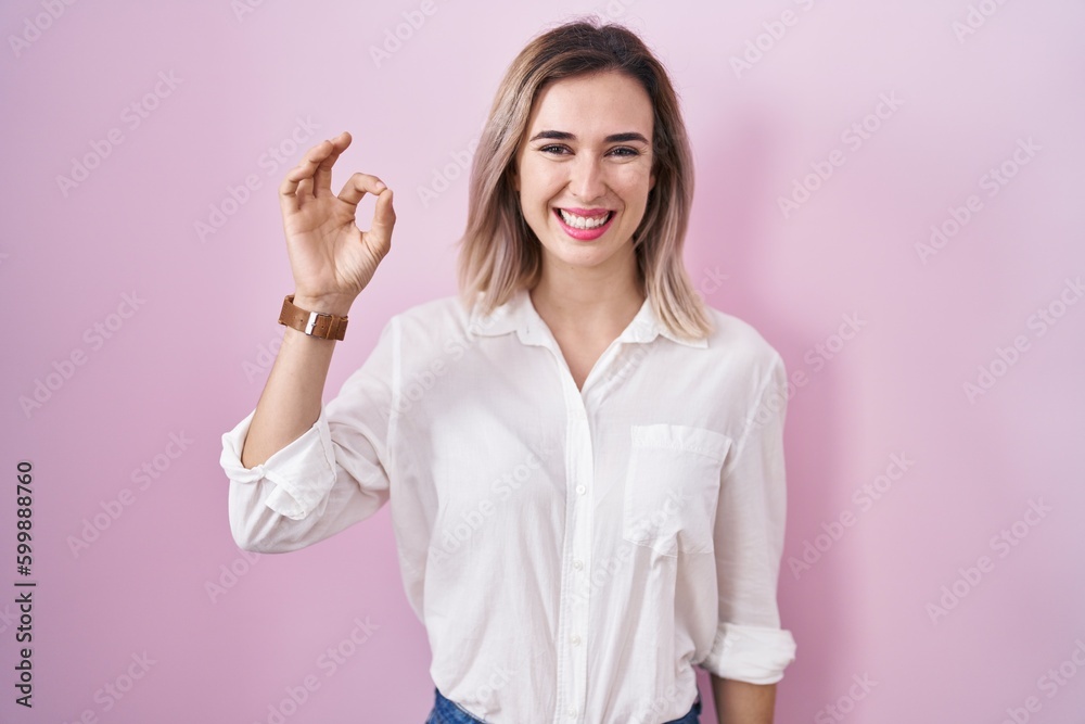 Young beautiful woman standing over pink background smiling positive doing ok sign with hand and fingers. successful expression. Young beautiful woman standing over pink background smiling positive doing ok sign with hand and fingers. successful expression.